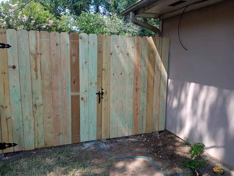 Wooden fence and gate with black hardware. Beige wall on right side. Green and brown wood.