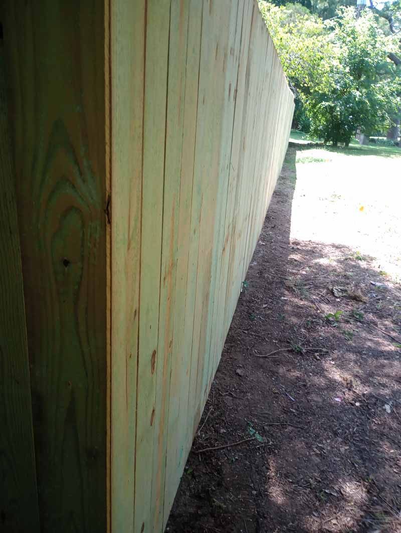Wooden fence in a natural setting. Brown dirt borders the fence. Green foliage in the background.
