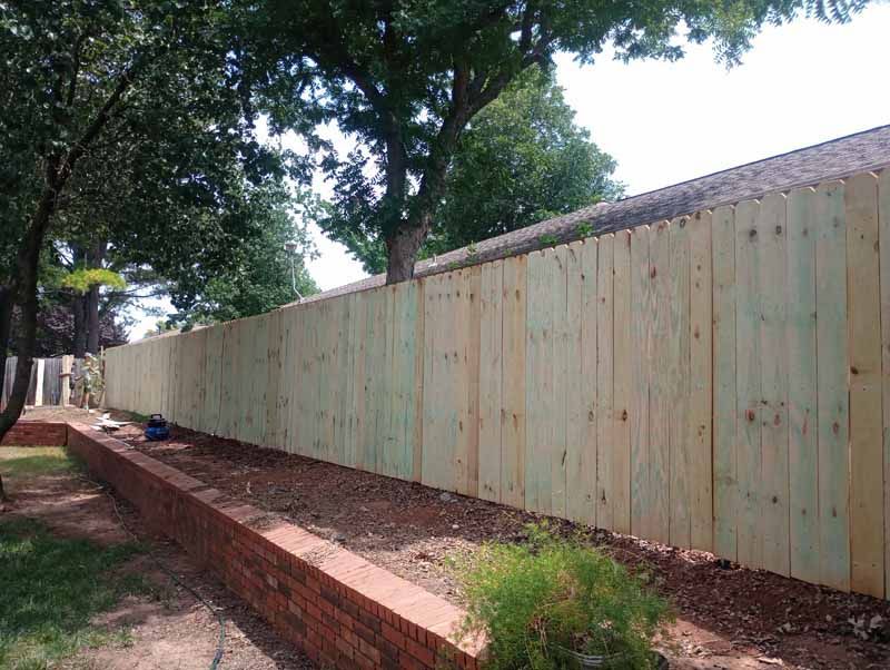 Wooden fence running along a brick planter bed. Green trees and clear sky in the background.