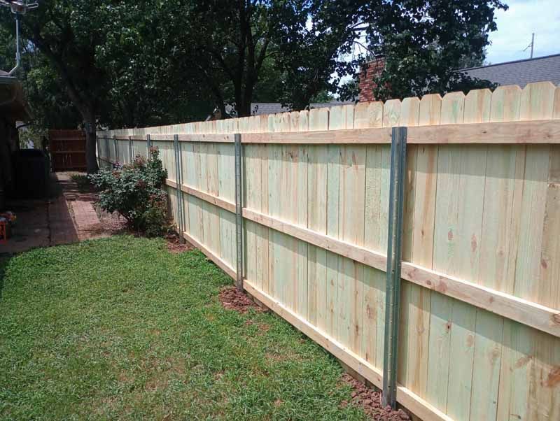 Wooden fence in a yard with green grass, steel posts, and trees in the background.