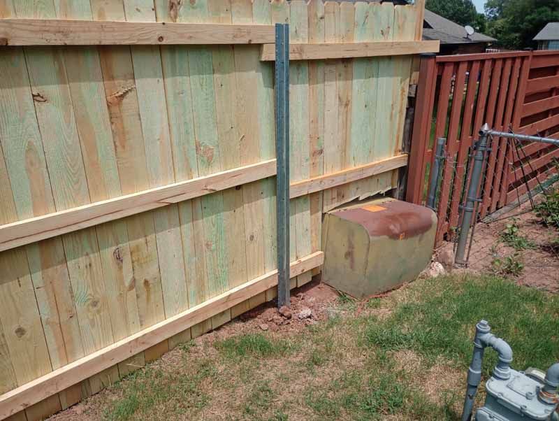 Wooden fence with a metal pole and a utility box in a grassy yard.
