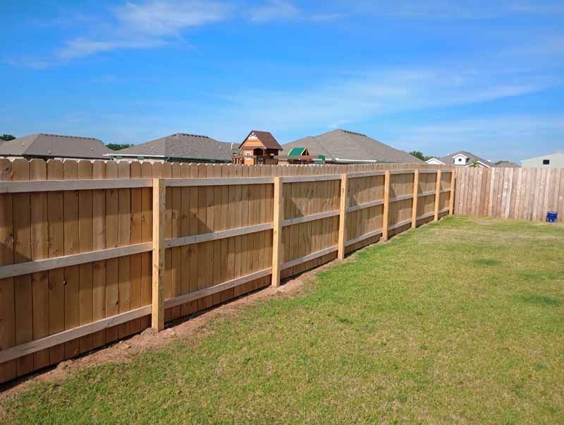 Wooden fence in a backyard with green grass and houses in the background under a blue sky.