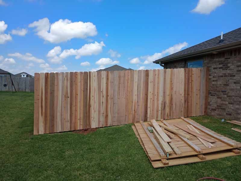 A newly built wooden fence in a backyard, partially blocking a brick house under a blue sky.