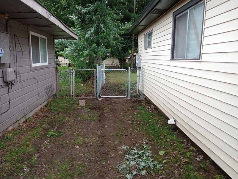 A chain-link fence and gate separate two houses; a grassy area lies between them.