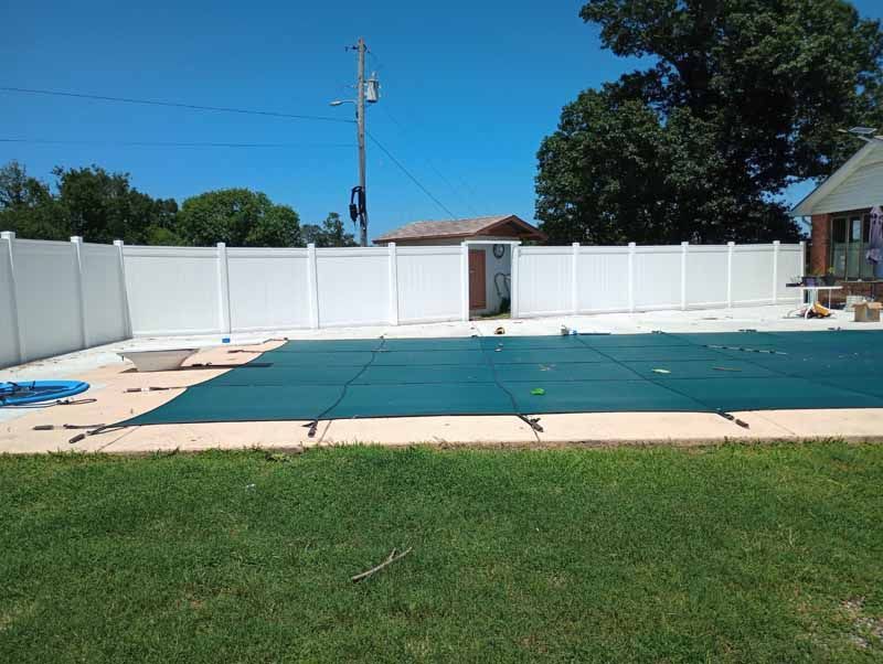 White fenced backyard with pool covered by a green tarp, on a sunny day.