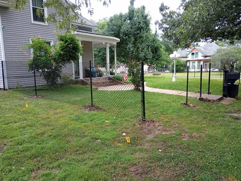 Chain-link fence in front yard of a house with a porch, lawn, trees, and walkway to the street.