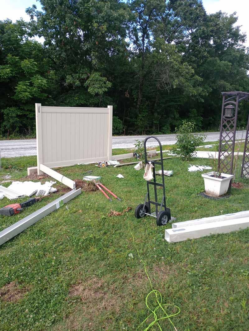 A partially built tan vinyl fence on a grassy lawn with tools and a hand truck.