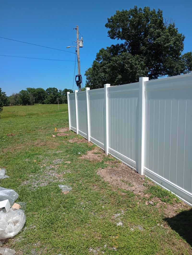 White vinyl fence in a grassy yard under a blue sky, with trees and power lines visible.
