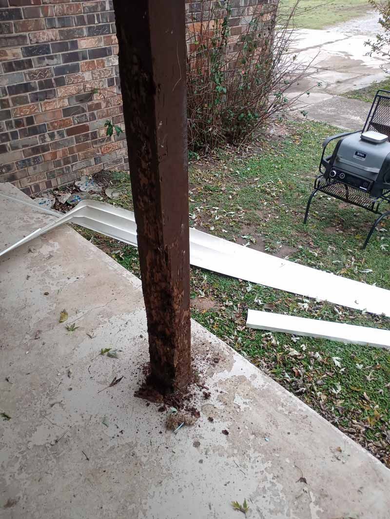 Brown, weathered porch support post on a concrete surface. White gutter pieces and yard in background.