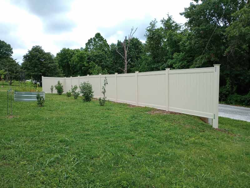 Beige vinyl fence on a grassy hill; trees in the background.