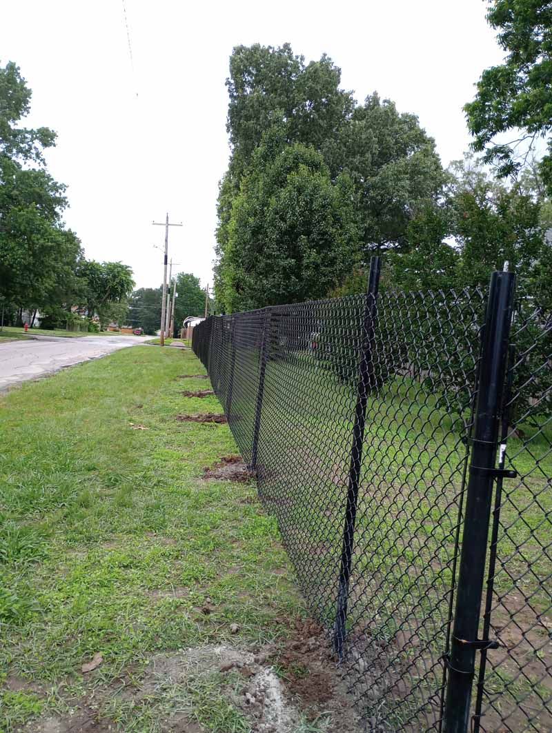 Black chain-link fence lines a grassy area beside a road, with trees and a utility pole visible.