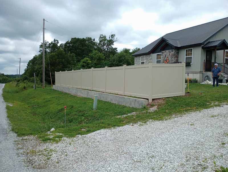 Beige fence along a concrete foundation, next to a gravel driveway and house on a cloudy day.