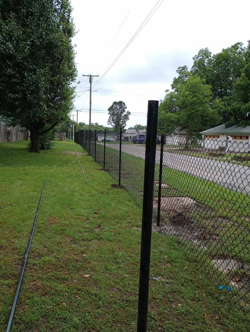 Black chain link fence along a grassy yard next to a street with houses and trees.