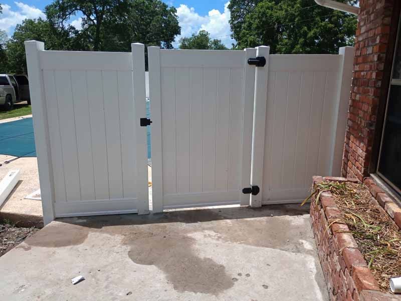White vinyl fence and gate next to a brick building on a concrete patio.