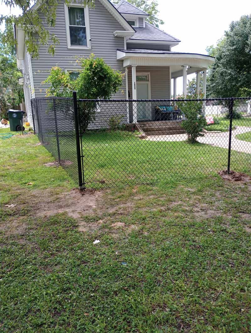 Black chain-link fence in front of a gray house with a porch and yard. Green grass and trees are visible.