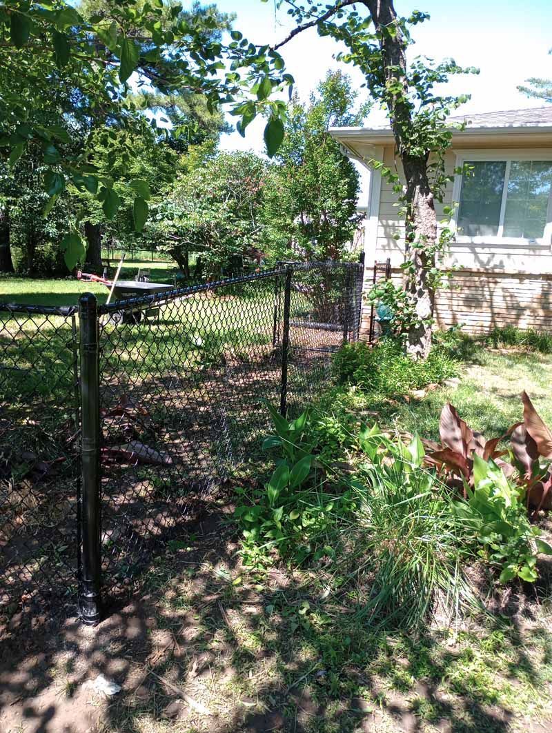 Black chain-link fence bordering a grassy yard next to a house with a tree.