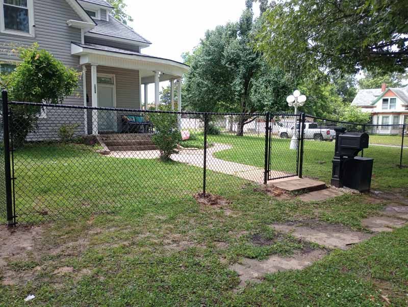 Black chain-link fence surrounds a house with a porch and yard, entrance through a gate, grass.