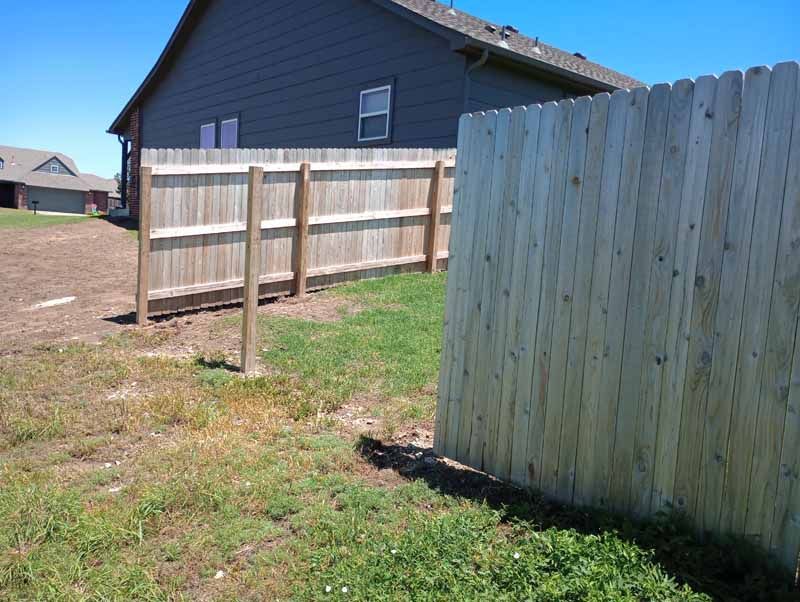 Wooden fence sections enclose a grassy backyard with a house in the background on a sunny day.