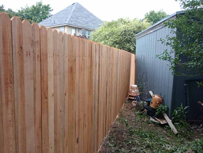 Wooden fence next to a gray shed in a yard, with a house visible in the background.