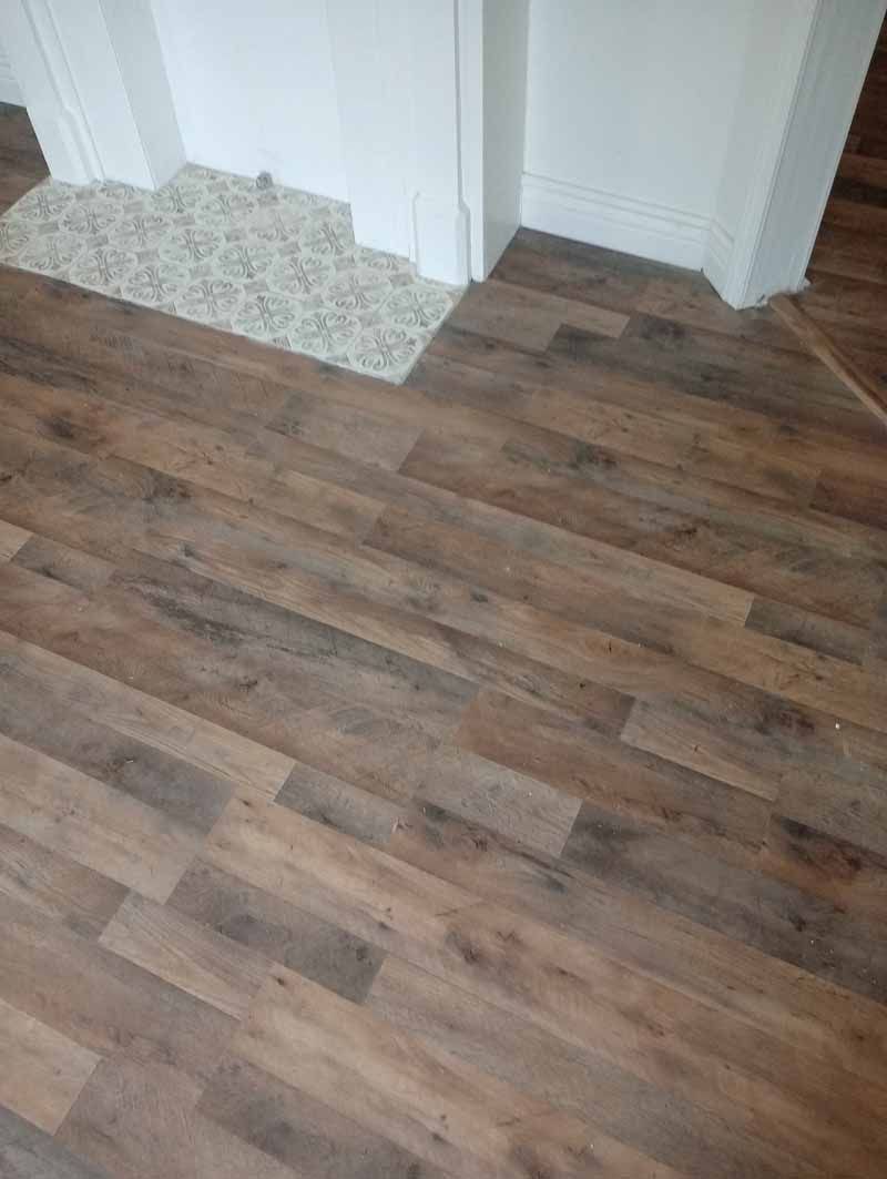 Wood-look flooring meets a doorway with a carpeted section. The floors are brown with light and dark streaks.