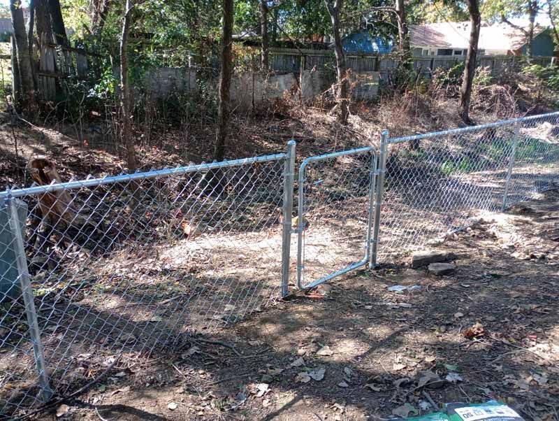 Chain-link fence with gate in wooded area; houses visible in background.