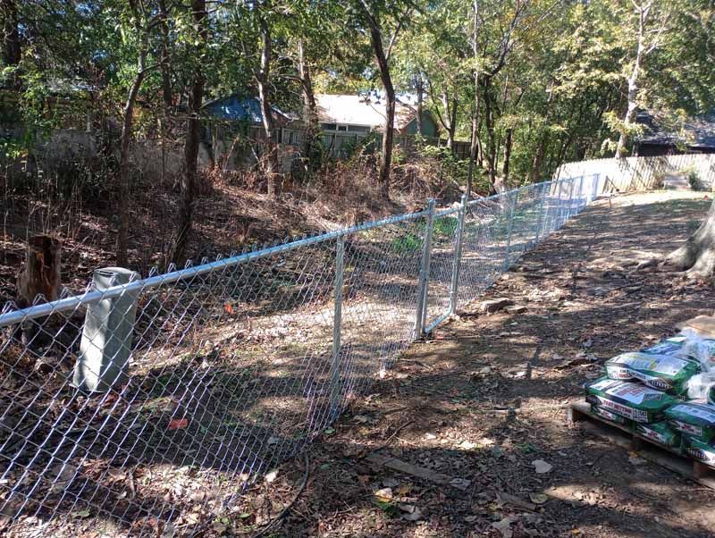 Chain-link fence in wooded area, light gray posts and mesh, bags of material on the right.
