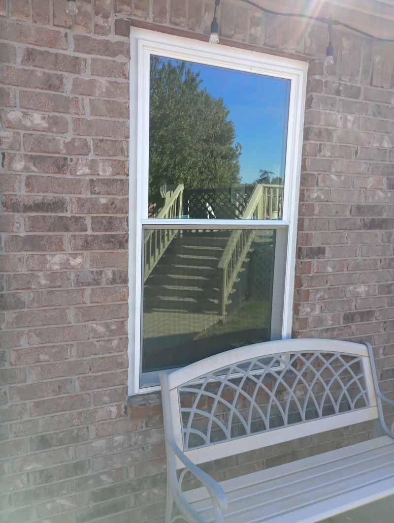 A tall window reflecting a tree, stairs, and sky, set in a brick wall above a white bench.