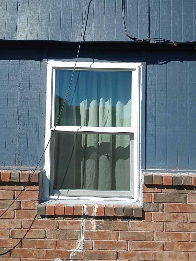 Window in a blue wall, with white frame and beige curtains, set above brick.
