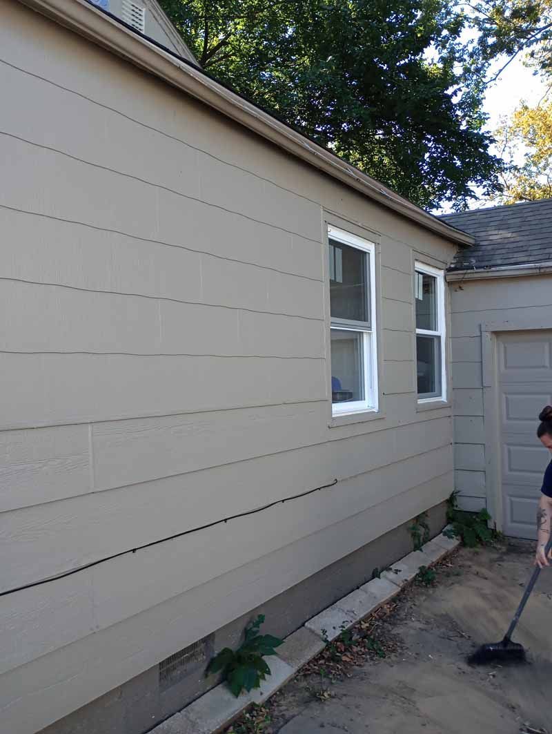 Side view of a beige house with two white-framed windows, a gutter, and a person sweeping the ground.