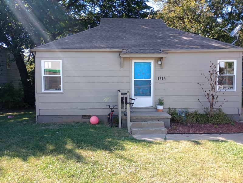 Small beige house with gray roof, front door with blue screen. Gray steps lead to the entrance. Grass lawn and trees surround.
