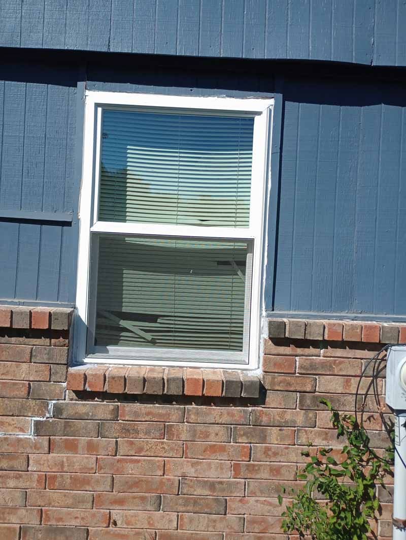 Window on a blue building with white trim, above a brick base, blinds closed.