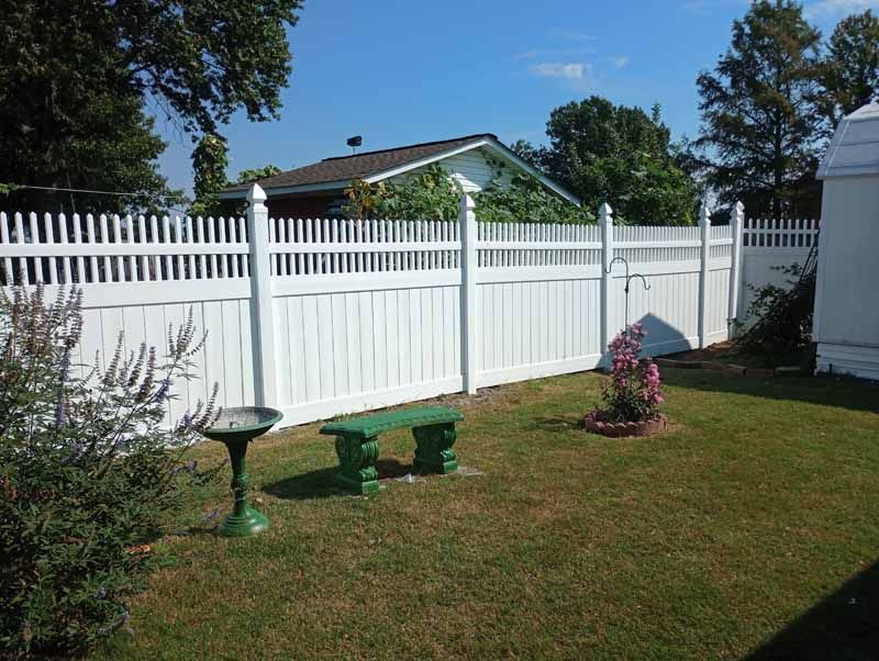 White picket fence surrounds a grassy yard with a green bench, birdbath, and roses.