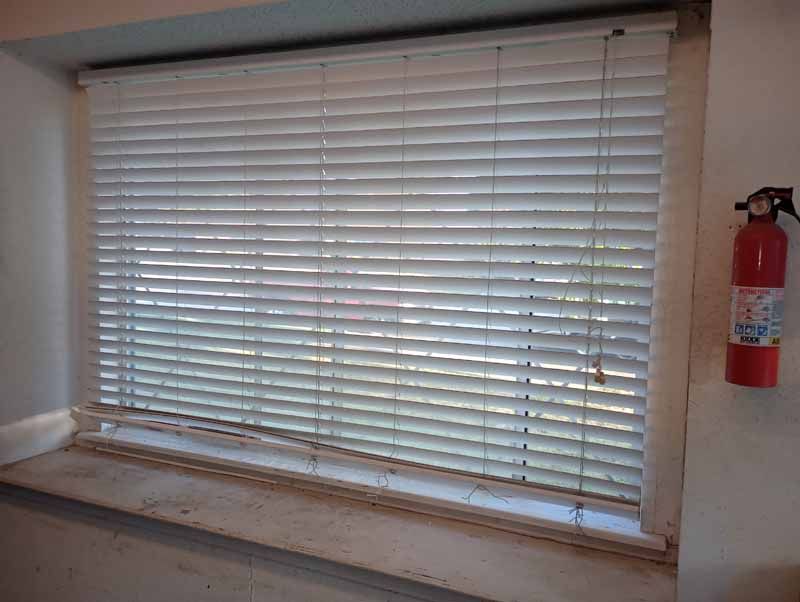 Window with white horizontal blinds, dusty sill, and red fire extinguisher on the wall.