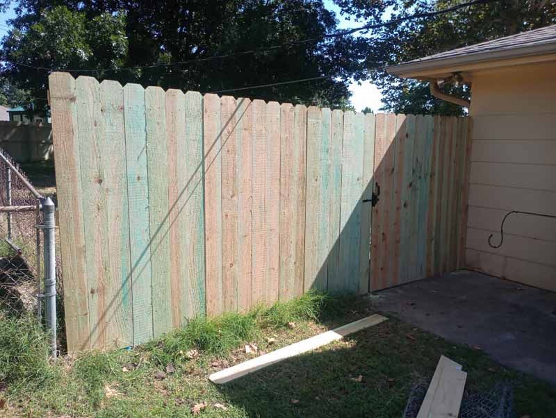 Wooden fence with a gate, green-tinted and new, next to a house with a concrete patio.