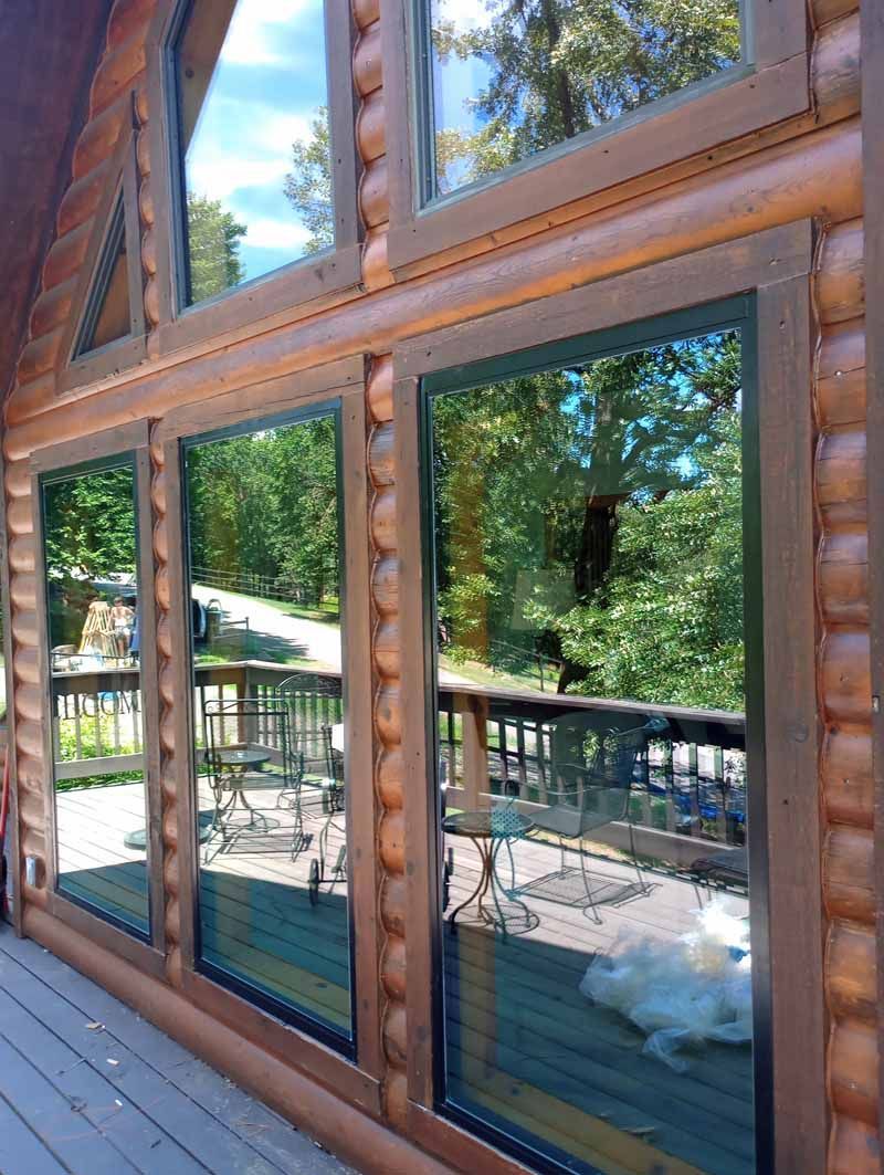 Large windows in a log cabin with a view of trees and a deck, reflecting the surroundings.