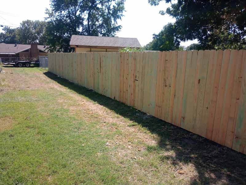 Wooden fence bordering a grassy backyard, with trees and houses visible in the background.