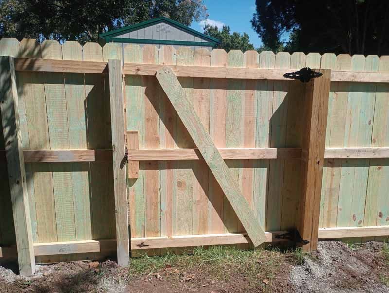Wooden fence gate, made of light-colored wood. Features a diagonal support beam and black hinges.