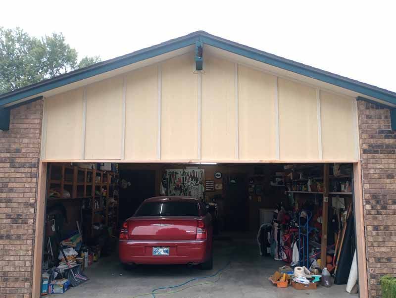 Red car parked in a cluttered garage, seen from the outside. Beige siding above the opening.