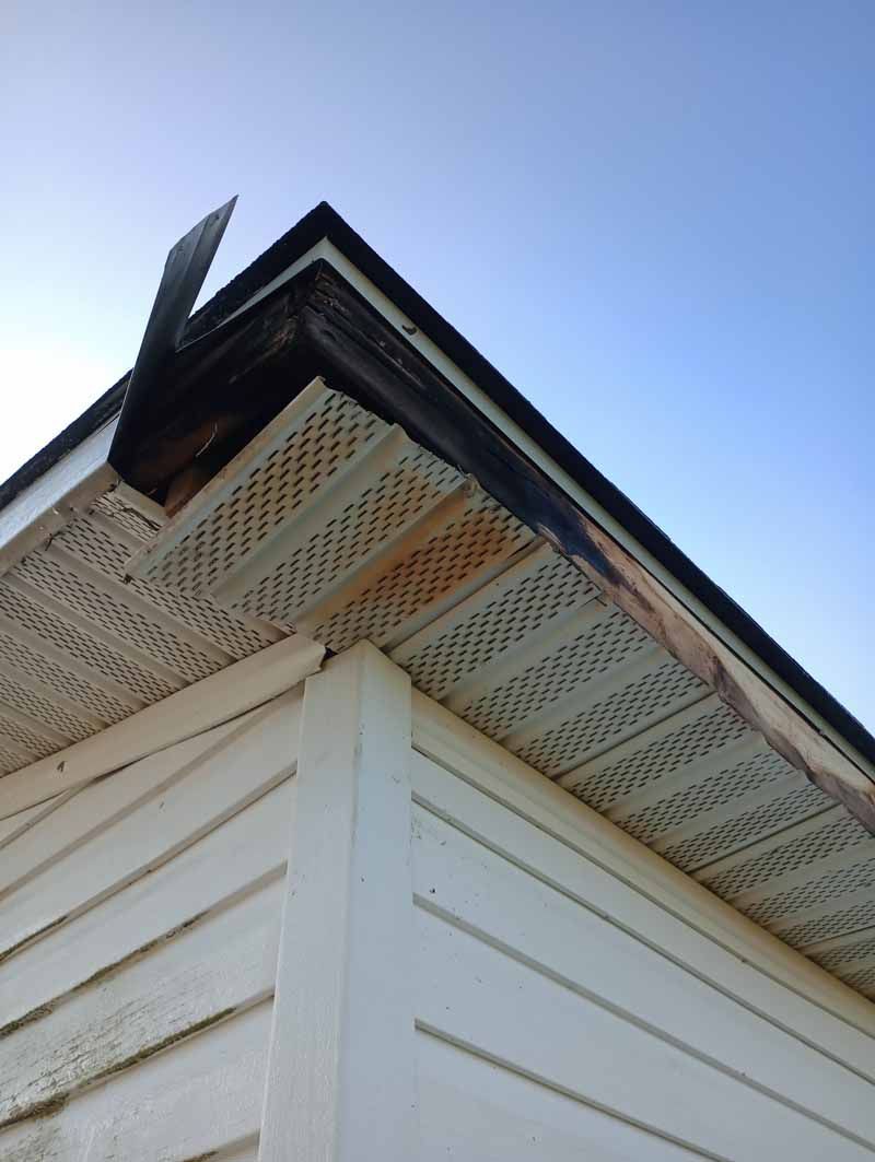 Corner of white house with damaged roof trim and vented soffit. Sky in background.