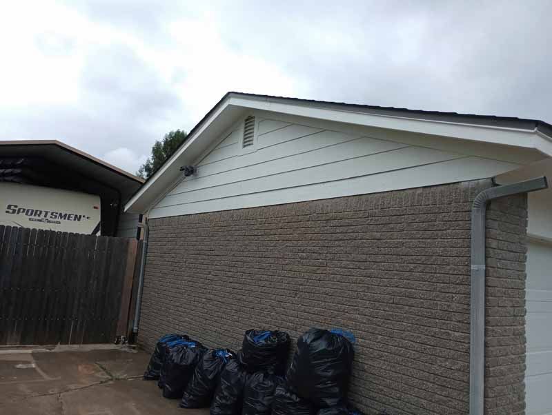 Garage with a brick facade and white siding under a cloudy sky. Bags of trash are on the ground.