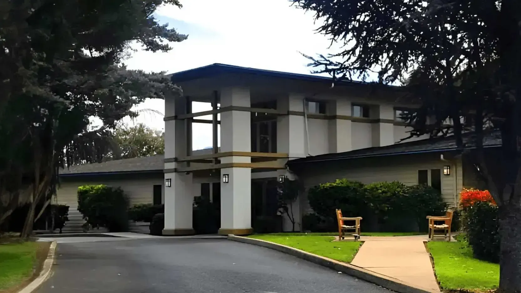 Cream-colored building with dark roof, surrounded by trees and green lawn; paved walkway.