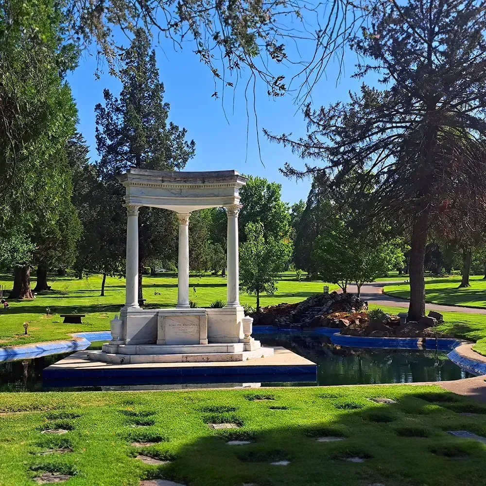 White marble gazebo with columns beside a blue-lined pond and green grass in a cemetery.