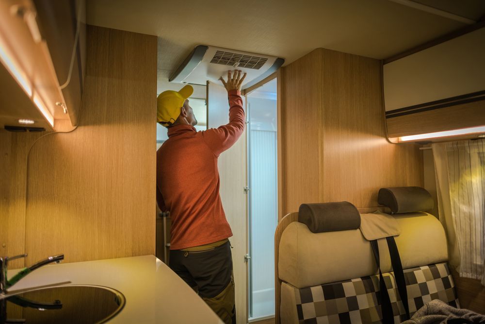 Man in orange shirt inside RV, adjusting air vent. Interior features light wood paneling, seating, and a sink — Lakeside Gas Fridges In Argenton, NSW