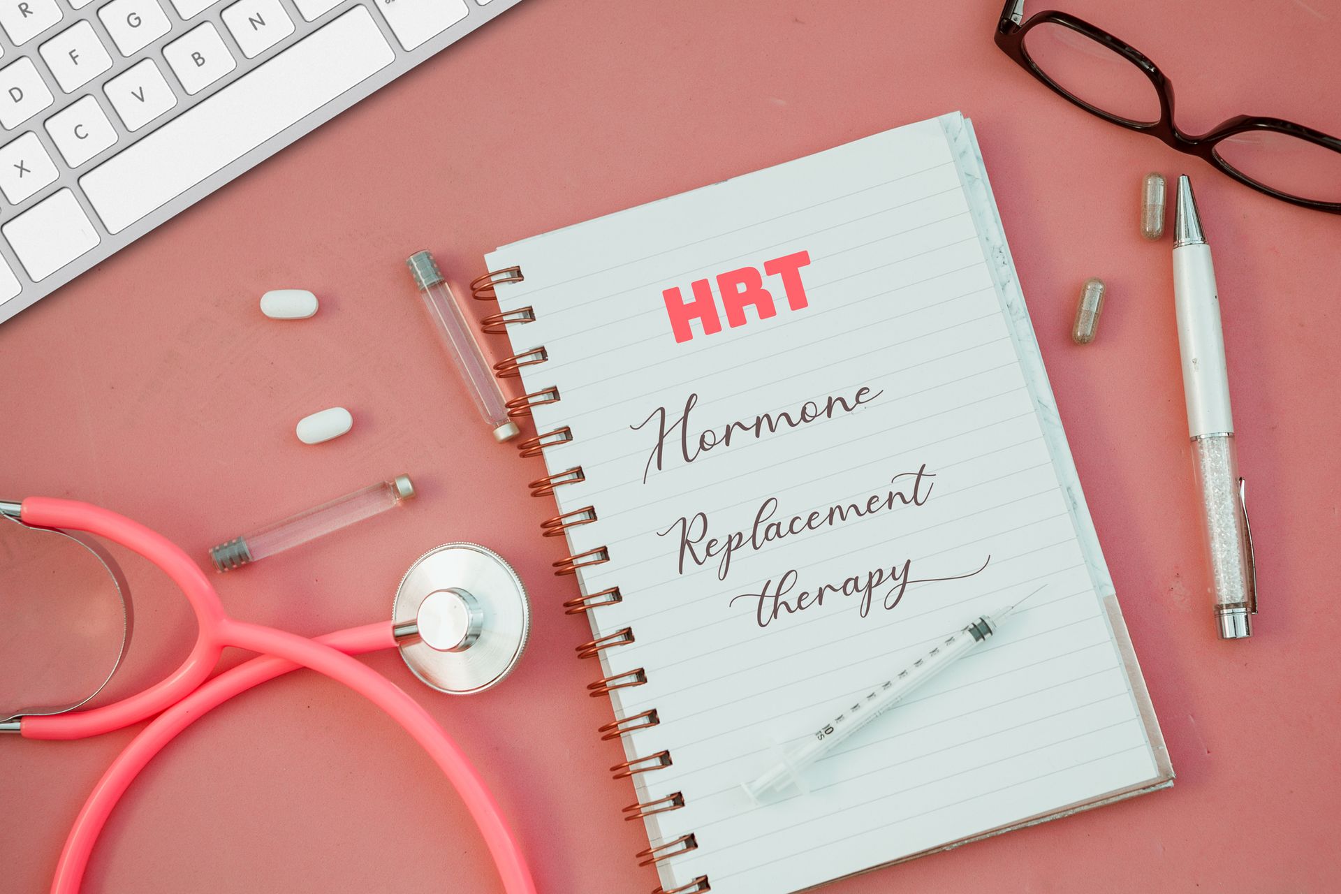 Female doctor’s desk: pink surface, keyboard, stethoscope, pills, open HRT notebook.