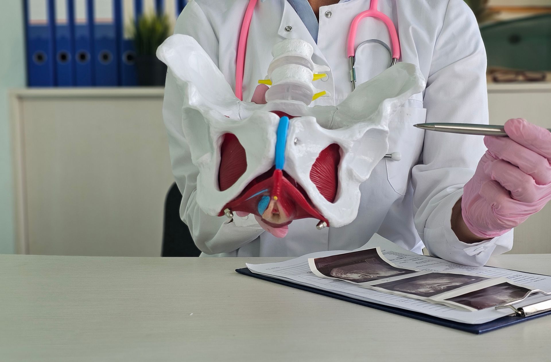 Doctor with stethoscope points to pelvic model, holding forceps; ultrasound scans on desk.