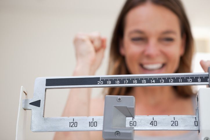 Closeup of weight scale on a number, with a blurry woman cheering on the background.