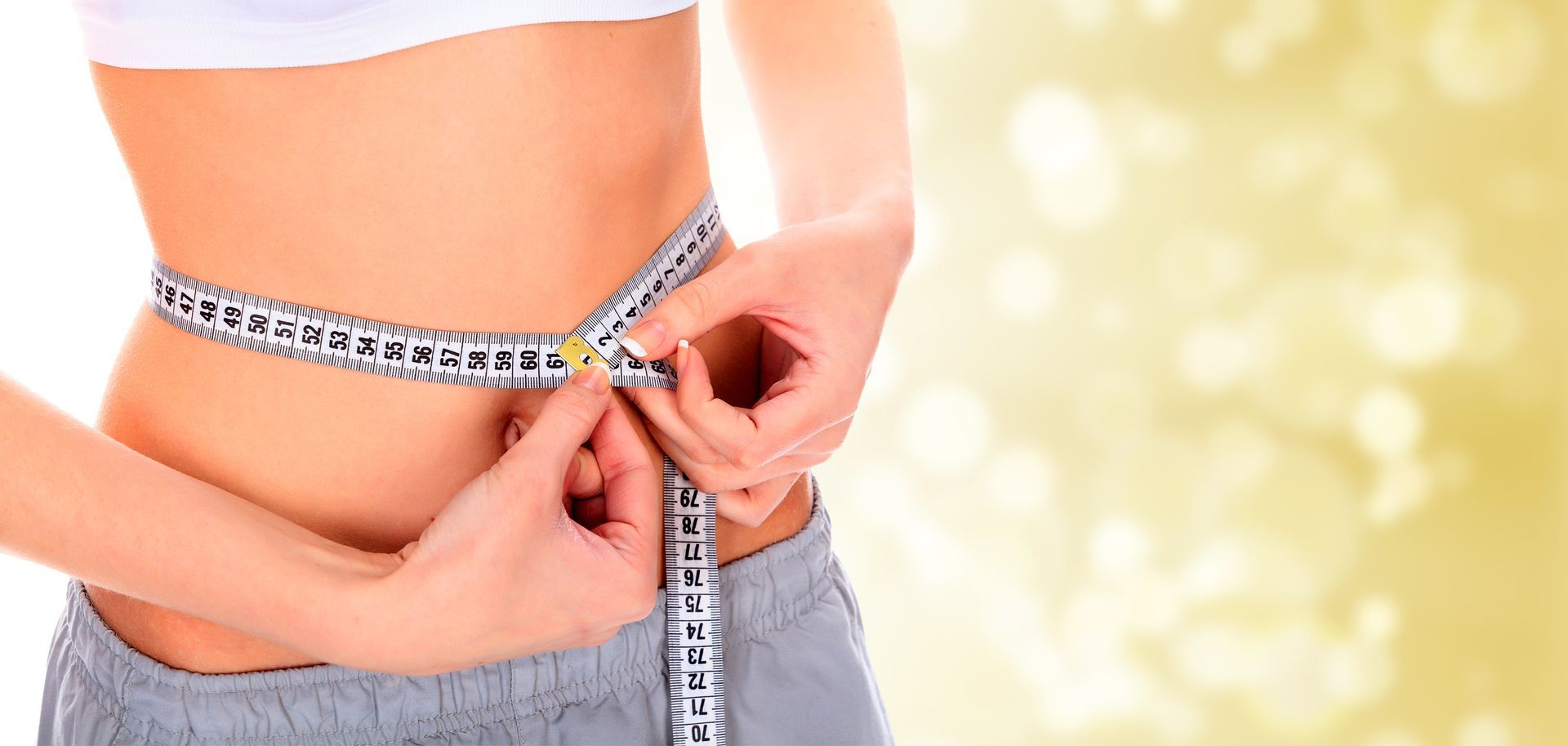 Closeup of woman measuring her waist with measuring tape, with a blurry bright background.