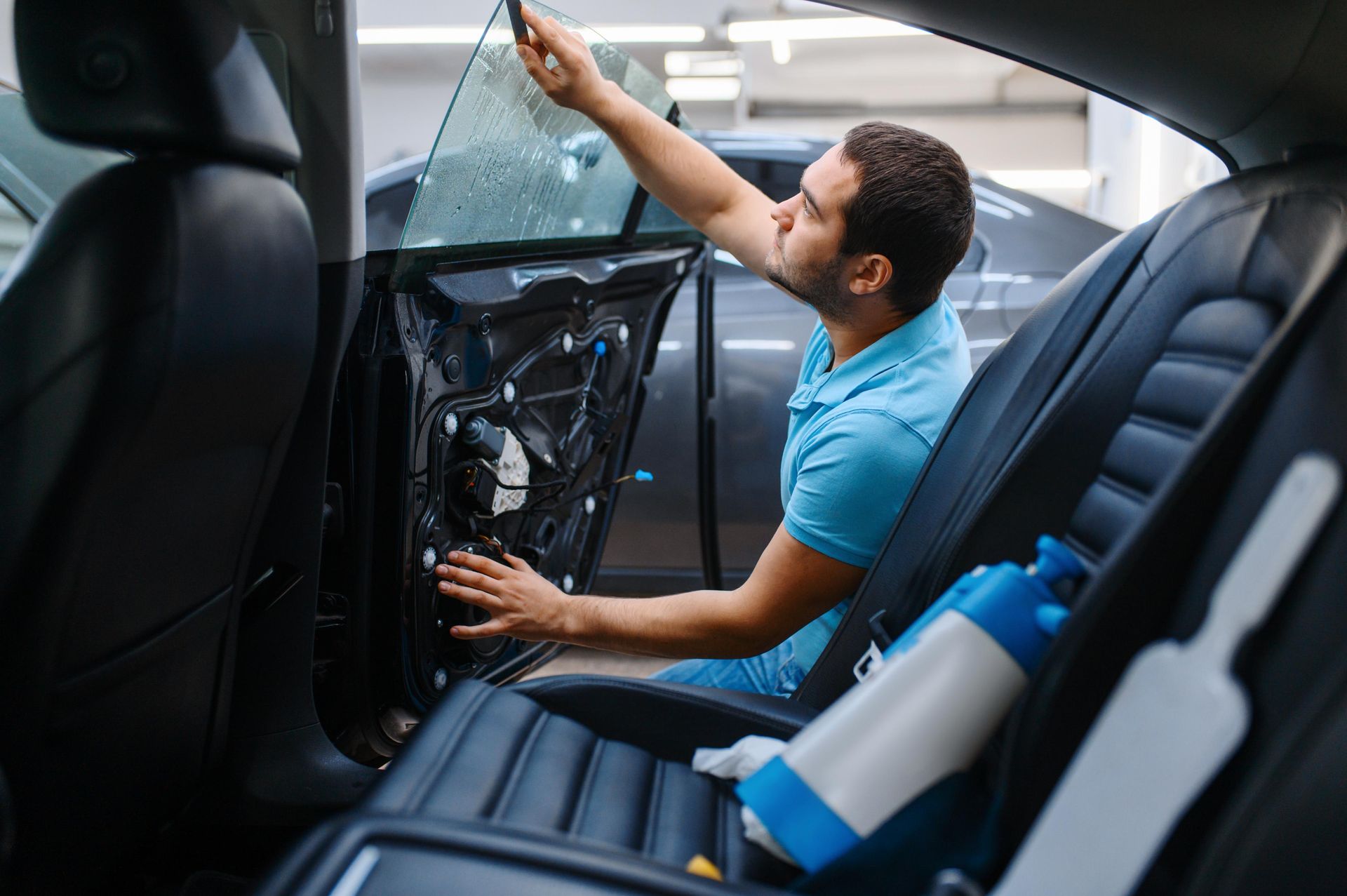 Man applying tint to a car window inside a car; blue shirt, spray bottles on seat.