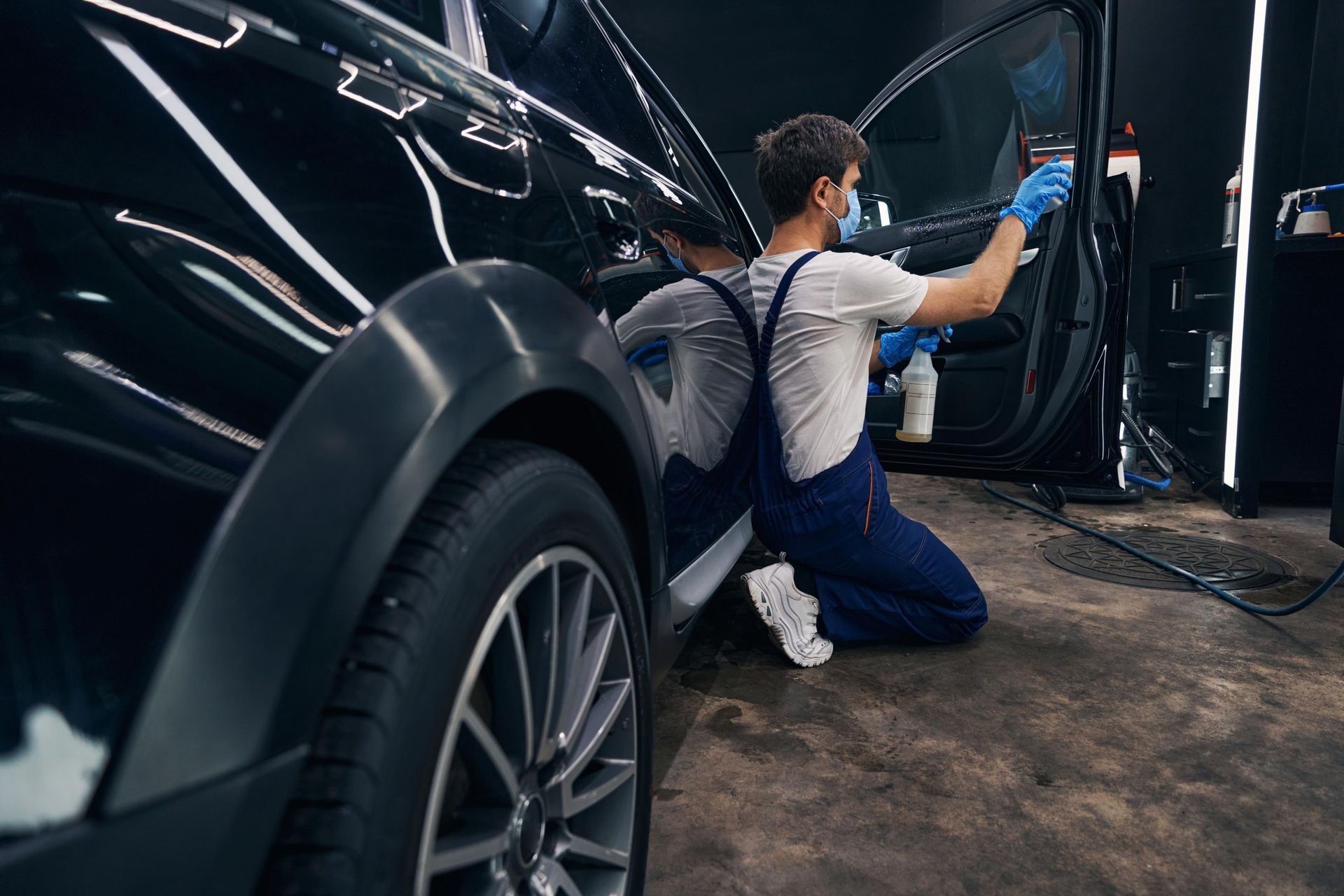 A person in blue overalls applies tint to a black car window inside a garage.