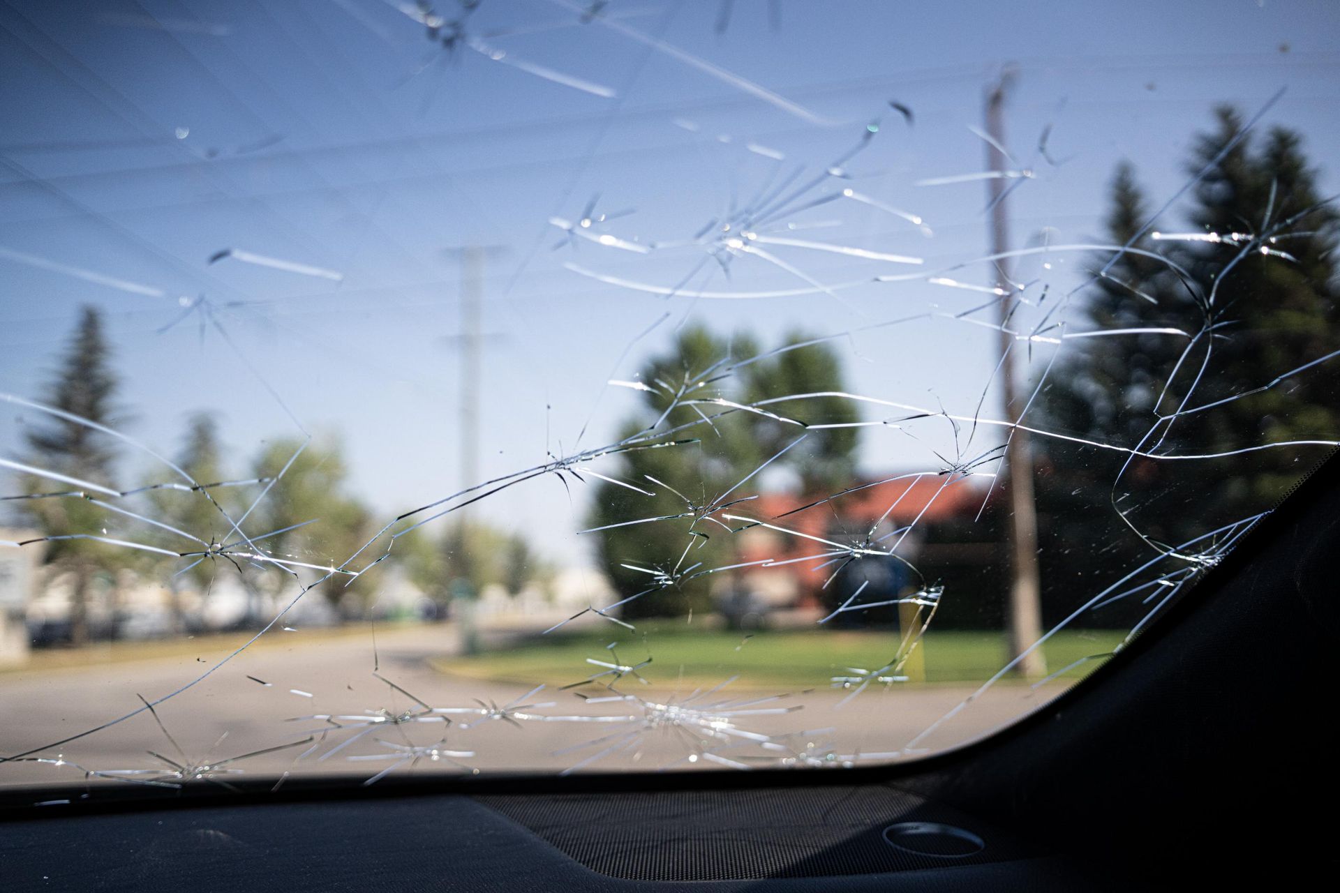 Cracked car windshield with a blurred view of trees, houses, and a blue sky in the background.